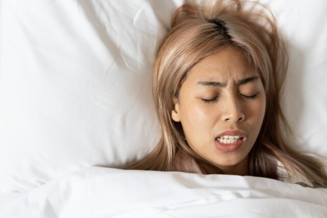 Woman with closed eyes and tense expression, clenching teeth in bed, illustrating stress-related bruxism during the holiday season.