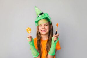 Girl in Halloween costume holding a lollipop and a toothbrush, promoting dental health during Halloween candy season.