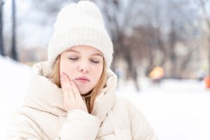 Young woman in winter attire, holding her jaw in discomfort, experiencing tooth sensitivity in cold weather.