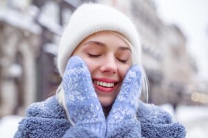 Young woman in winter attire smiling and protecting her face from cold, illustrating the importance of dental care during winter to manage tooth sensitivity.