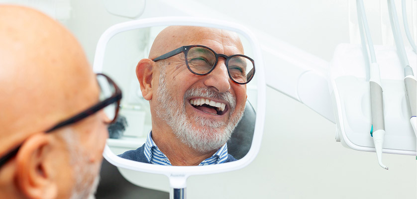 Smiling older man with glasses looking into a dental mirror, reflecting satisfaction after dental treatment, illustrating positive dental implant experience.