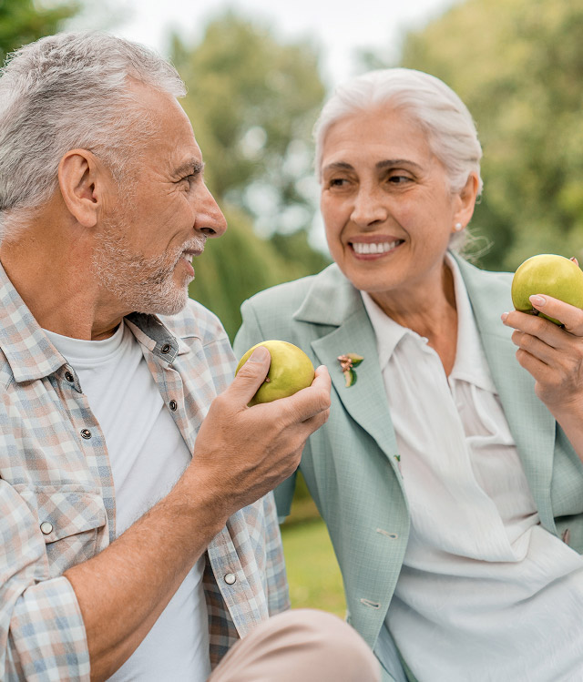 Older couple enjoying green apples outdoors, smiling and engaging in conversation, highlighting healthy lifestyle choices relevant to dental health.